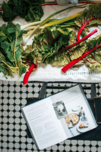 Preparations for Zelnik Pie with the cookbook splayed open on a black bar stool next to a table of leafy green ingredients