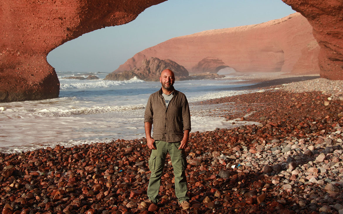 Man stands, hands in pocket, on a rocky beach with the ocean and wind shaped sand arches in the background.